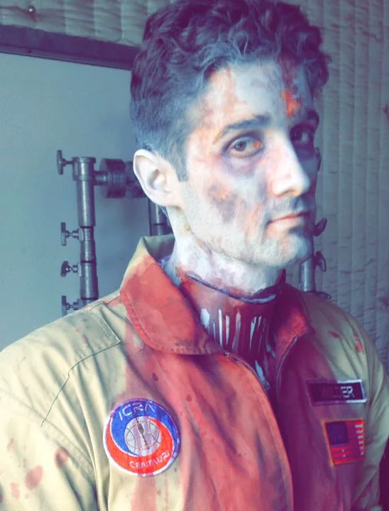 Fictional special effects makeup showing facial wounds on a performer in a work jumpsuit.